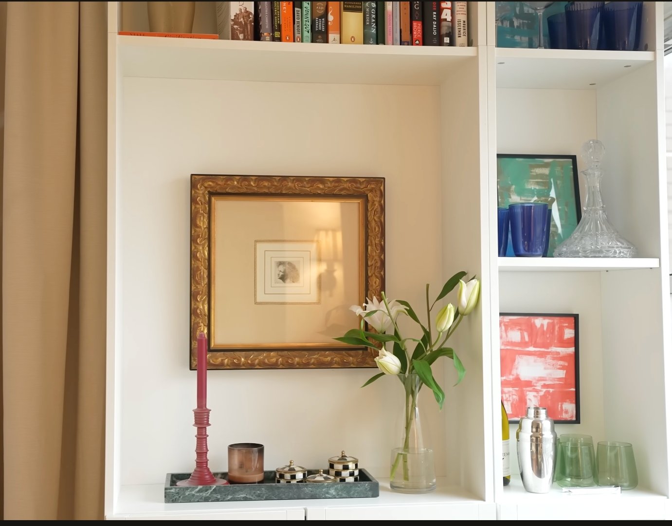 Curated bookshelf with gilt frame, marble tray, lilies, and colorful art