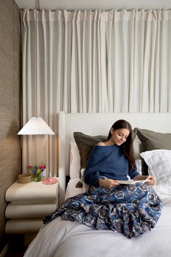 Matilde Cascella reading on a designed bed with pleated lamp, draped curtains, and grasscloth walls