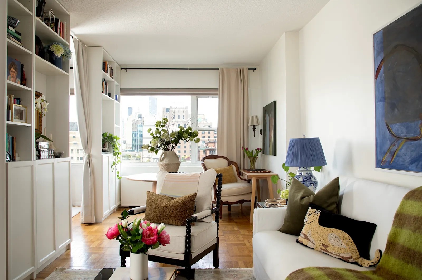 Light-filled NYC living room with white bookshelves, blue abstract art, chinoiserie lamp, and city views